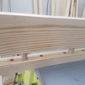 May include: Close-up of a wooden shelf with dowel pins and holes for assembly. The shelf is made of unfinished pine wood.