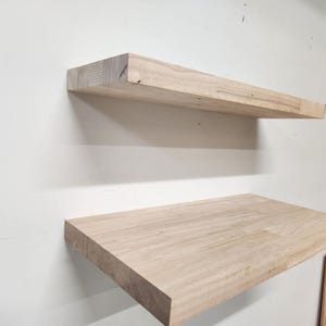 May include: Two unfinished, light-colored wooden floating shelves mounted on a white wall. The top shelf is rectangular and extends from the wall. The bottom shelf is also rectangular and is positioned below the top shelf.
