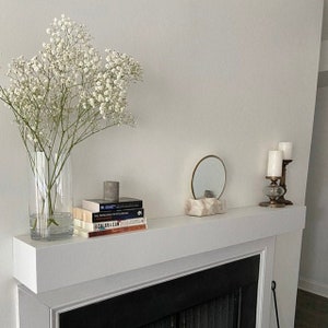 May include: A white fireplace mantel with a stack of books, a small round mirror, and a candle holder with two candles. A bouquet of white baby's breath flowers is in a clear glass vase on the mantel.