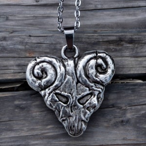 May include: A silver pendant necklace with a ram skull design. The pendant has a detailed, textured surface and is suspended from a matching chain. The necklace is displayed against a rustic wooden background.