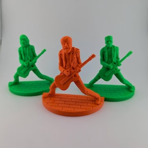 May include: Three brightly coloured figurines of guitarists. The central figure is orange, standing on a brick-patterned base, with the other two green figures on green bases. Each figure is holding a guitar.
