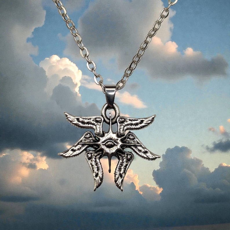 May include: A silver-toned pendant necklace featuring a detailed, stylized design with multiple wings and an eye at the center. The pendant hangs from a chain against a cloudy sky background. The necklace is a piece of jewelry.