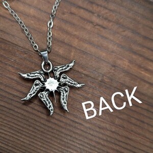 May include: A silver-colored pendant necklace featuring a stylized sunburst design. The pendant has a central circular element with radiating, wing-like shapes. The necklace chain is also silver. The word "BACK" is printed in white.