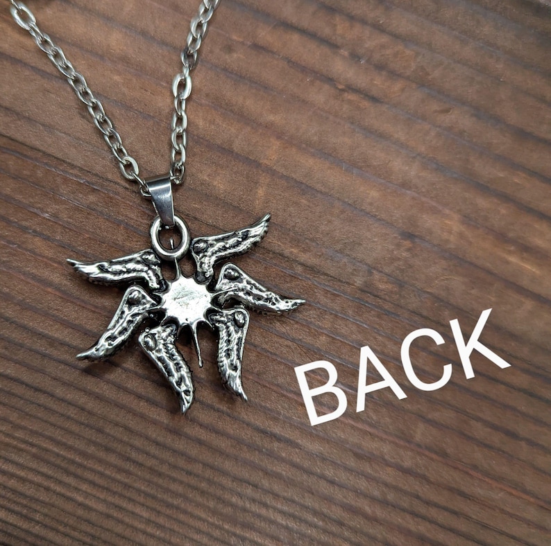 May include: A silver-colored pendant necklace featuring a stylized sunburst design. The pendant has a central circular element with radiating, wing-like shapes. The necklace chain is also silver. The word "BACK" is printed in white.