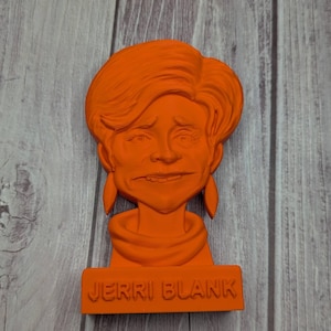 May include: Orange bust of a person with the name "JERRI BLANK" at the base. The bust has detailed facial features and hair. The background is a wooden surface.