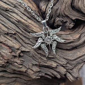 May include: A silver-colored pendant necklace featuring a stylized winged design. The pendant has multiple wings and a central eye-like detail. The necklace chain is also silver and hangs against a textured wooden background.