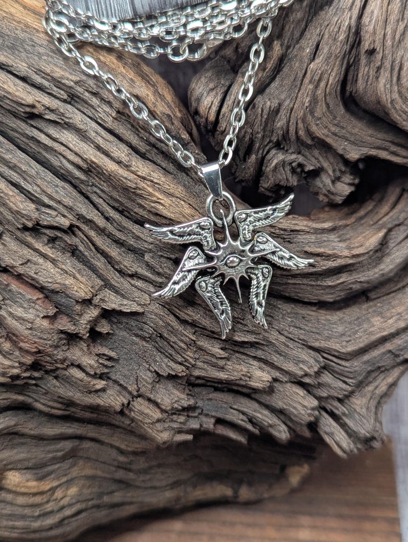 May include: A silver-colored pendant necklace featuring a stylized winged design. The pendant has multiple wings and a central eye-like detail. The necklace chain is also silver and hangs against a textured wooden background.