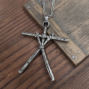 May include: A silver necklace with a pendant shaped like a stick figure made of sticks. The pendant is tied together with rope. The necklace is on a wooden surface.