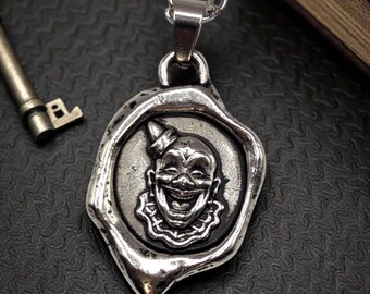 Puede incluir: Collar con colgante plateado que presenta una cara de payaso sonriente dentro de un diseño de sello de cera. El colgante cuelga de una cadena. En el fondo hay una llave y un libro.