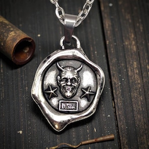 May include: Silver-toned pendant necklace featuring a devil face design with horns, two stars, and the text "MYSTIC SEER". The pendant is attached to a silver chain. The necklace is displayed on a dark wooden surface.