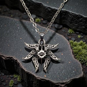 May include: Silver-toned pendant necklace featuring a detailed, symmetrical design with multiple wings and an eye in the center. The pendant hangs from a delicate chain. The necklace is displayed on a dark, textured surface.