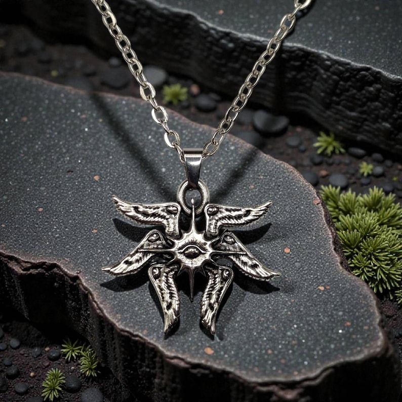 May include: Silver-toned pendant necklace featuring a detailed, symmetrical design with multiple wings and an eye in the center. The pendant hangs from a delicate chain. The necklace is displayed on a dark, textured surface.