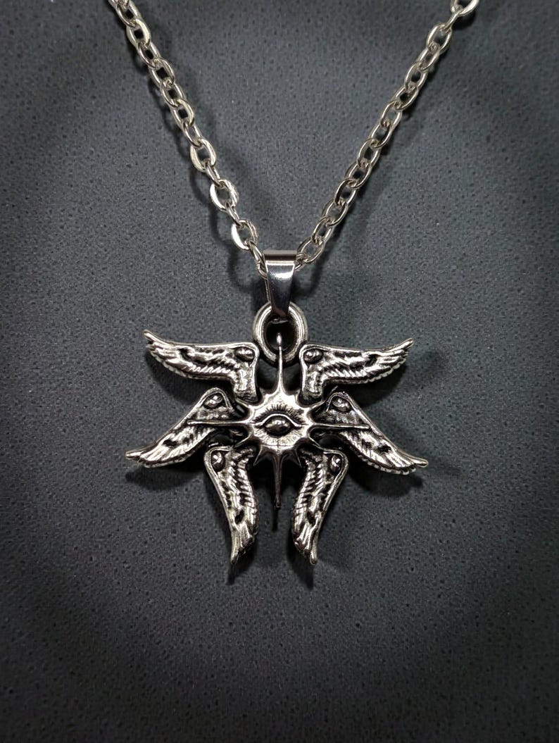May include: Silver-toned pendant necklace featuring a detailed, stylized design of wings and an eye. The pendant hangs from a delicate chain. The intricate design is visible against a dark background.