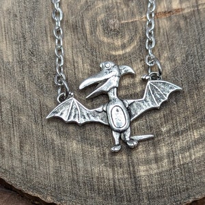 May include: Silver necklace with a pterodactyl pendant. The pendant is detailed with wings and a long beak.