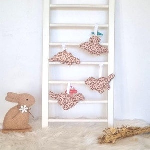 Puede incluir: Una escalera de madera blanca exhibe cuatro adornos de dinosaurios con estampado de leopardo y cintas blancas. Un conejito de peluche con una flor está sobre una alfombra blanca y esponjosa. Flores secas en la parte inferior derecha.