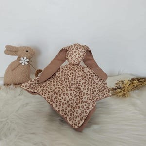 May include: A soft, plush baby security blanket with a bunny head and long ears. The blanket features a tan and brown leopard print pattern. A second stuffed bunny is in the background, with a white flower attached.
