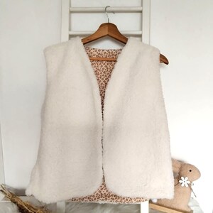 May include: A white faux fur vest with a leopard print lining. The vest is hanging on a wooden hanger.