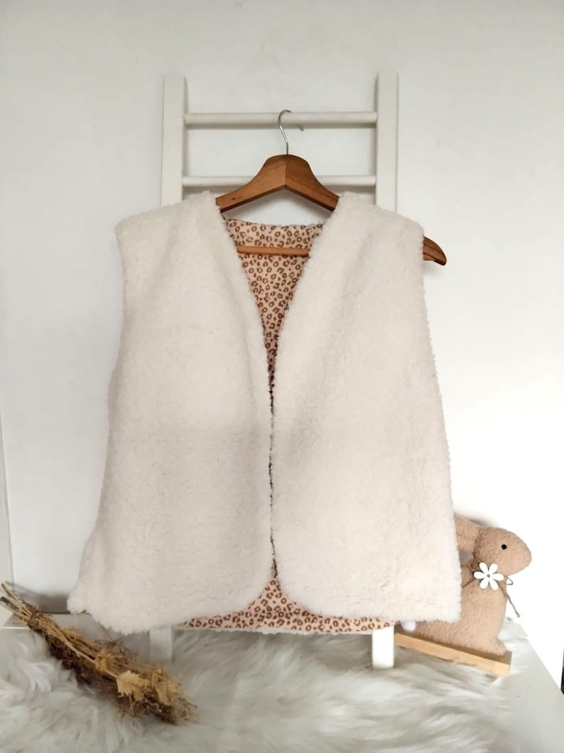 May include: A white faux fur vest with a leopard print lining. The vest is hanging on a wooden hanger.