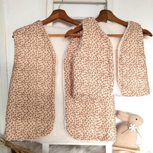 May include: Two brown and beige patterned fleece vests with a sherpa lining hanging on wooden hangers. The vests are on a white background.