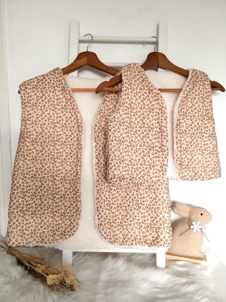 May include: Two brown and beige patterned fleece vests with a sherpa lining hanging on wooden hangers. The vests are on a white background.