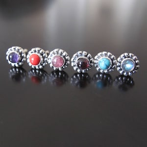 Puede incluir: Una fila de seis pendientes de botón con piedras preciosas de colores. Cada pendiente presenta un engaste plateado con cuentas alrededor de una piedra central. Las piedras son moradas, rojas, rosas, rojo oscuro, turquesas y celeste.