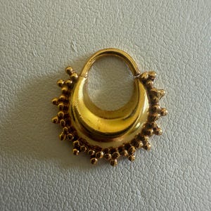 Puede incluir: Un pendiente dorado con un diseño en forma de media luna. El pendiente presenta un cierre circular y está adornado con pequeños adornos esféricos alrededor del borde exterior. El diseño general es ornamentado y detallado, con un acabado pulido.