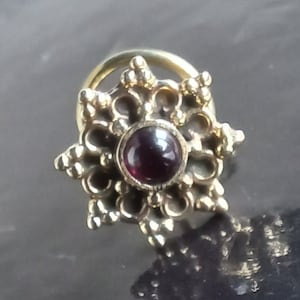 Peut inclure: Un anneau de nez en or avec un motif floral décoratif et une pierre précieuse violette foncée au centre.