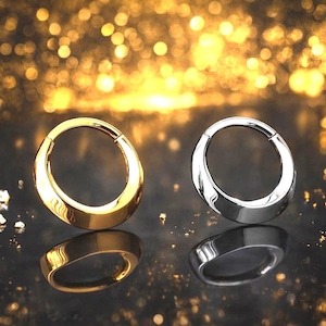 May include: Two circular body jewelry pieces. One is gold-colored, the other is silver-colored. Both have a smooth, polished finish and a hinged design. The background is a dark surface with golden bokeh.