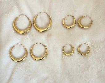May include: Four pairs of polished gold-tone hoop earrings arranged in two rows. The left column features two pairs of larger, thicker hoops, while the right column displays two pairs of smaller, thinner hoops, all with a smooth, reflective finish.