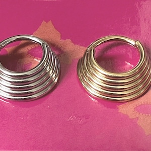 Könnte beinhalten: Zwei dekorative Ohrringe, einer silberfarben und einer goldfarben. Jeder hat ein kreisförmiges Design mit mehreren gestapelten, gebogenen Bändern. Die Ohrringe werden auf einer rosa Oberfläche präsentiert.