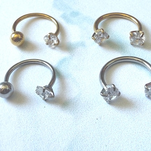 Peut inclure: Quatre bijoux de corps en forme de fer à cheval. Deux sont dorés avec une boule et une gemme claire à facettes. Les deux autres sont argentés, l'un avec une boule et l'autre avec deux gemmes claires à facettes. Le tout sur une surface bleu clair.