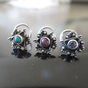 May include: Three silver nose rings with different colored gemstones. The rings are in the shape of a star with a triangle in the center. The gemstones are turquoise, pink, and blue.