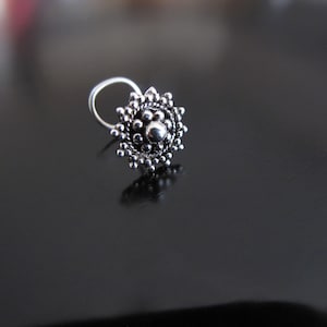 May include: A silver nose ring with a round, ornate design featuring a central bead and a circular pattern of smaller beads.