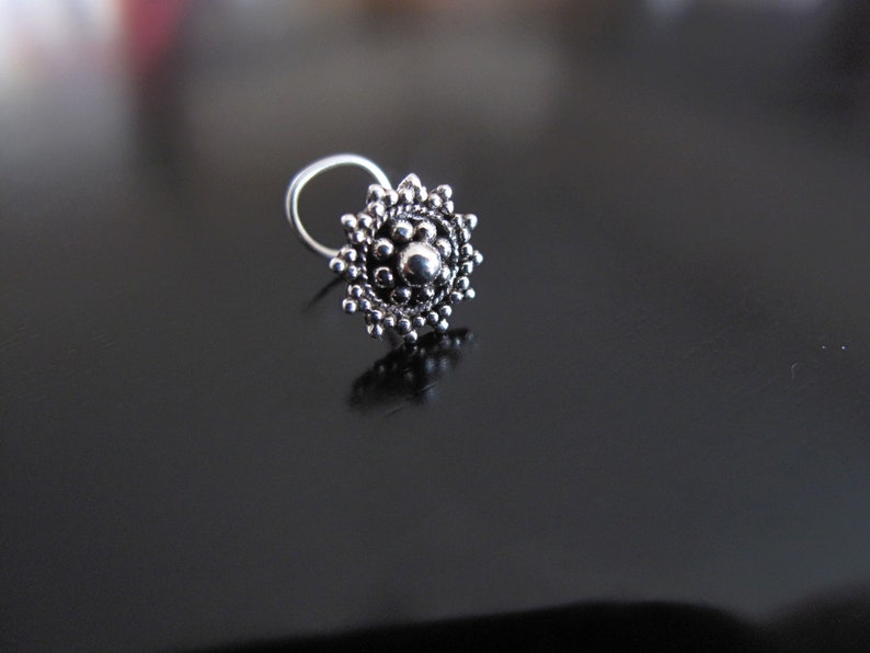May include: A silver nose ring with a round, ornate design featuring a central bead and a circular pattern of smaller beads.