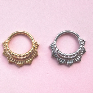 Può includere: Due anelli per septum decorativi, uno dorato e uno argentato. Ogni anello presenta un motivo floreale con una fila di perline. Lo sfondo è rosa chiaro uniforme. Gioielli per il corpo.