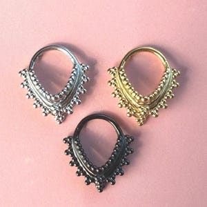 Könnte beinhalten: Drei dekorative Septumringe in Silber, Gold und Schwarz. Jeder Ring hat ein Perlendesign am unteren Rand, das ein strukturiertes, ornamentales Aussehen erzeugt. Die Ringe werden auf einem rosa Hintergrund präsentiert.