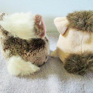 May include: Two plush animal toys, seen from behind, are placed next to each other. One is brown and white, the other is tan with brown details. Both have fluffy tails and ears, and are made of soft, plush material.