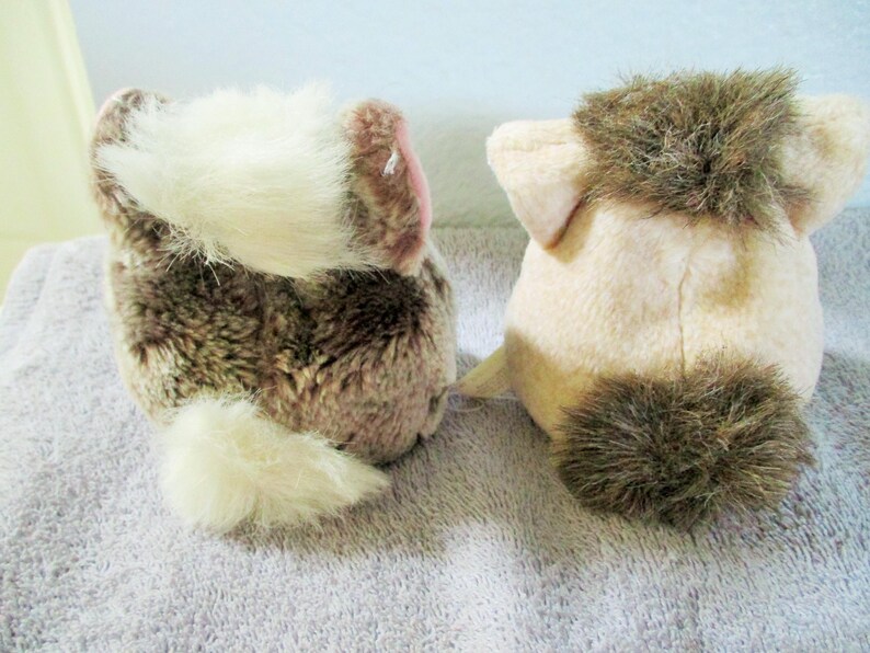 May include: Two plush animal toys, seen from behind, are placed next to each other. One is brown and white, the other is tan with brown details. Both have fluffy tails and ears, and are made of soft, plush material.