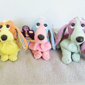 May include: Three plush Hush Puppies dog toys in orange, blue, and purple. Each has floppy ears, a black nose, and a tag. The orange dog has yellow accents, the blue dog has pink accents, and the purple dog has green accents. They are sitting on a light gray surface.