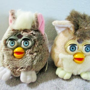May include: Two vintage Furby toys. One is brown with pink accents, the other is tan with brown accents. Both have blue eyes, yellow beaks, and red mouths. They are plush and have white feet.