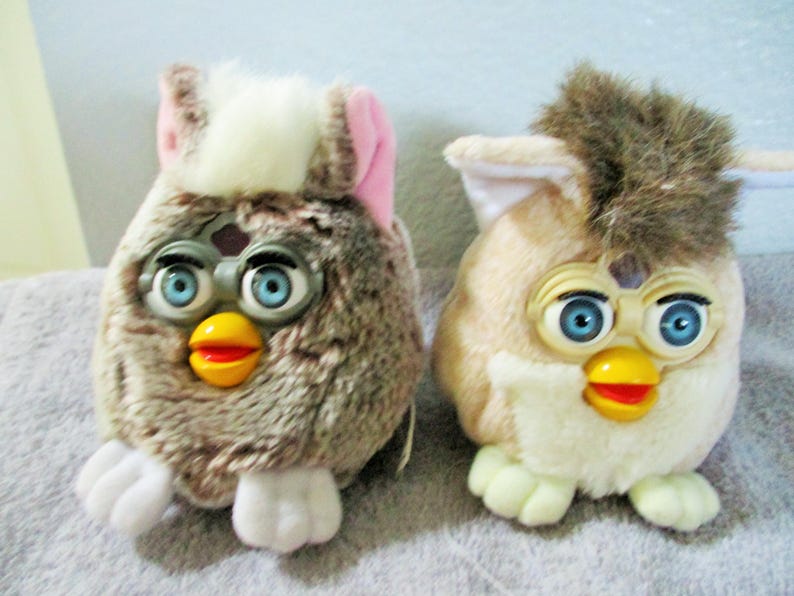 May include: Two vintage Furby toys. One is brown with pink accents, the other is tan with brown accents. Both have blue eyes, yellow beaks, and red mouths. They are plush and have white feet.