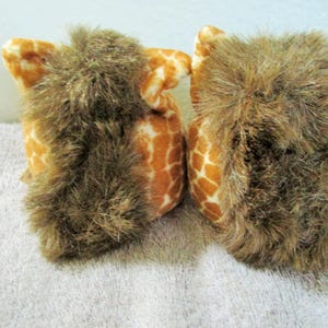 May include: Two plush owl toys. The owls have a giraffe-like pattern in shades of orange and white. The owls have brown, shaggy fur around their faces. The toys are sitting on a white surface.