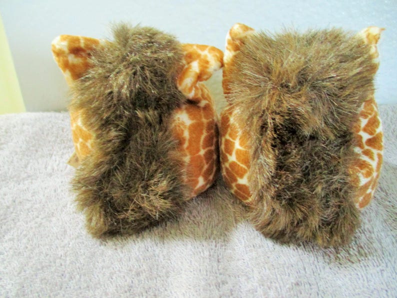 May include: Two plush owl toys. The owls have a giraffe-like pattern in shades of orange and white. The owls have brown, shaggy fur around their faces. The toys are sitting on a white surface.