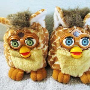 May include: Two vintage Furby toys. Each has a giraffe-like pattern in shades of brown and tan, with a yellow beak and red mouth. One has green eyes, the other blue.