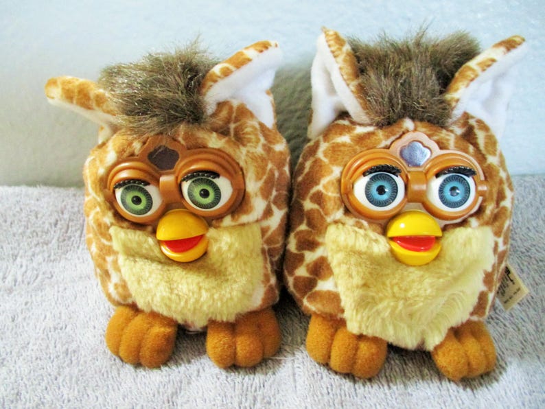 May include: Two vintage Furby toys. Each has a giraffe-like pattern in shades of brown and tan, with a yellow beak and red mouth. One has green eyes, the other blue.