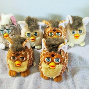 May include: A group of six vintage Furby toys in various colours and patterns. The Furbies feature large eyes, yellow beaks, and soft fur. The designs include brown, tan, and patterned styles, evoking a sense of nostalgia.