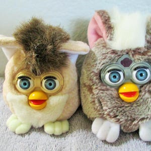 May include: Two vintage Furby toys. One is tan with brown accents, the other is brown with pink and white accents. Both have blue eyes, yellow beaks, and white feet. They are plush and have a retro aesthetic.
