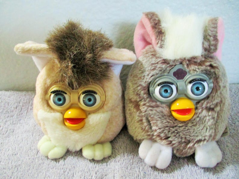 May include: Two vintage Furby toys. One is tan with brown accents, the other is brown with pink and white accents. Both have blue eyes, yellow beaks, and white feet. They are plush and have a retro aesthetic.