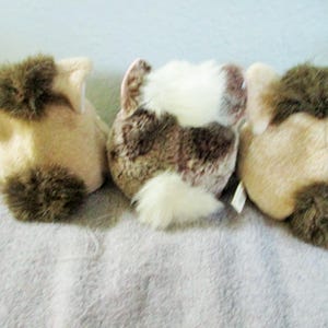 May include: Three plush animal toys, seen from behind. They are beige and brown, with fluffy brown tails and accents. The central toy has a white and brown pattern. The toys are on a light grey surface.