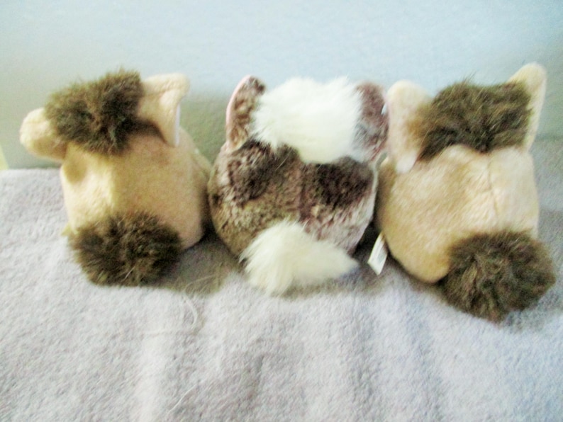 May include: Three plush animal toys, seen from behind. They are beige and brown, with fluffy brown tails and accents. The central toy has a white and brown pattern. The toys are on a light grey surface.